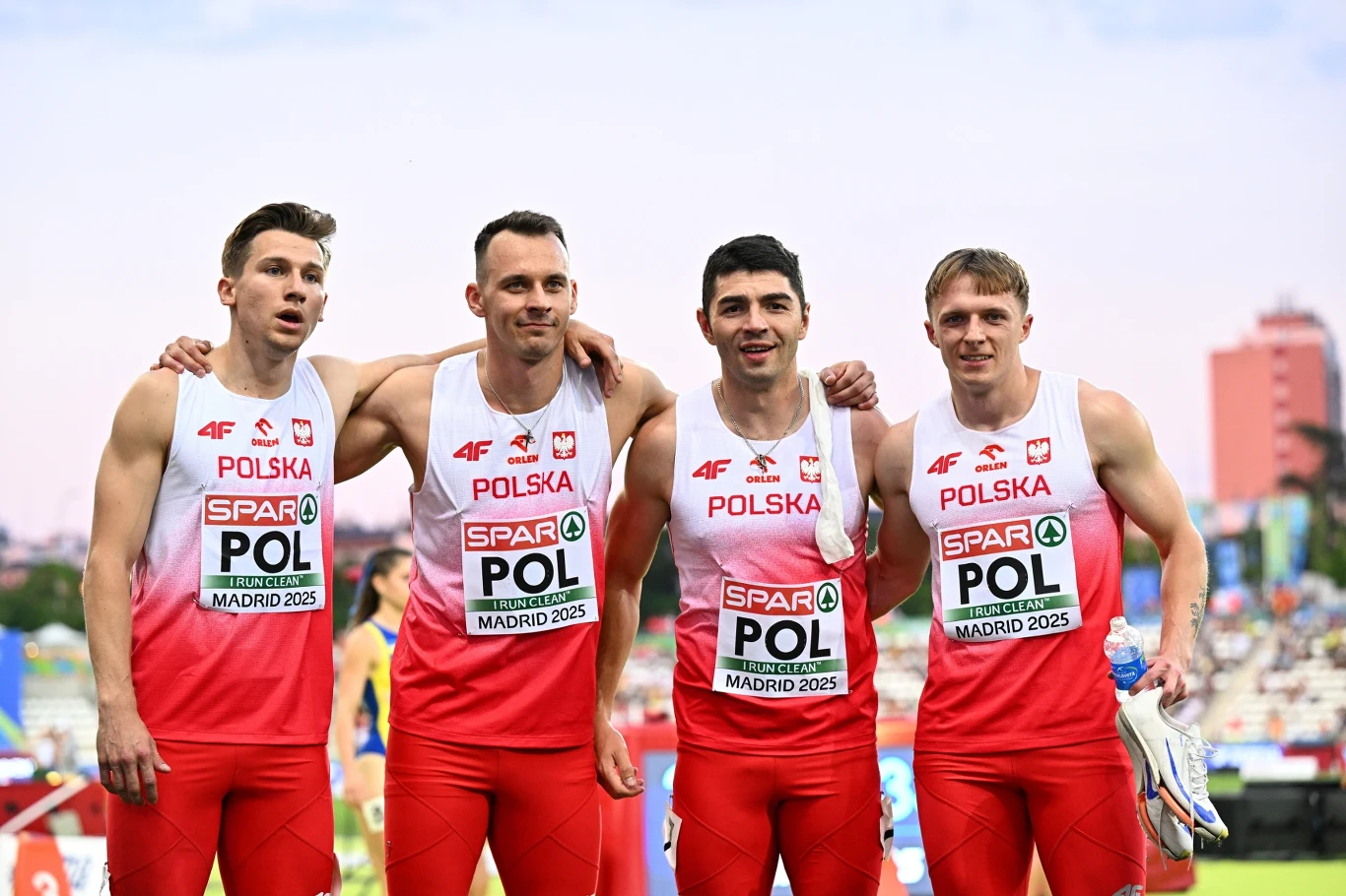 Czterech lekkoatletów reprezentacji Polski w czerwono-białych strojach sportowych z napisem POLSKA na piersi, stojących ramię w ramię na stadionie lekkoatletycznym po zakończonym biegu sztafetowym, w tle widoczni kibice i fragmenty miejskiego krajobrazu.