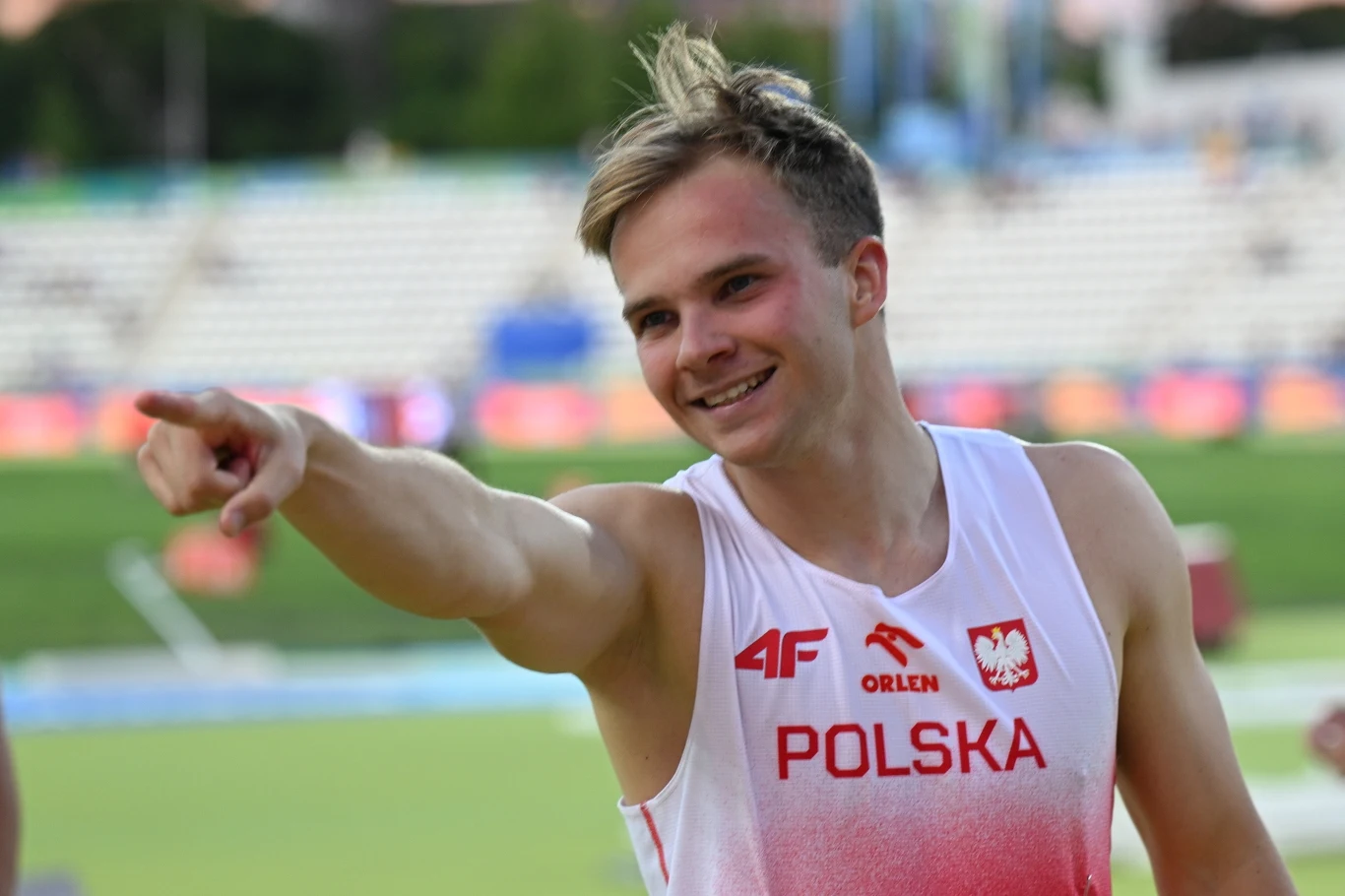 Uśmiechnięty młody sportowiec w stroju reprezentacji Polski z napisem 'POLSKA' wskazuje ręką w bok, stojąc na stadionie lekkoatletycznym w otoczeniu rozmytego tła trybun.