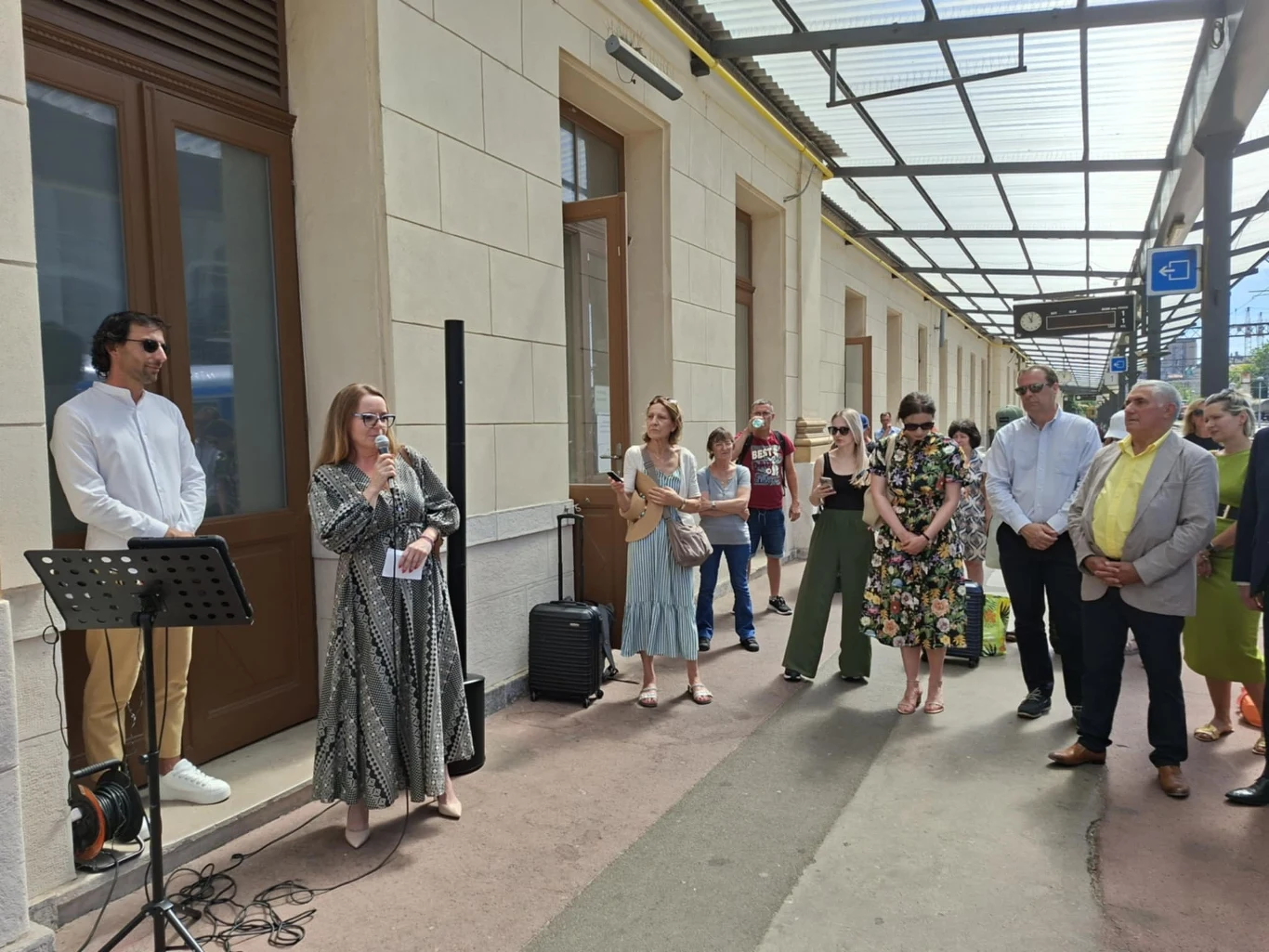 Grupa ludzi zgromadzona pod zadaszonym chodnikiem obok budynku, jedna kobieta przemawia przez mikrofon, reszta uczestników słucha. Po lewej stronie stoi mężczyzna przy statywie z nutami, a w tle widoczna jest elewacja i okna.