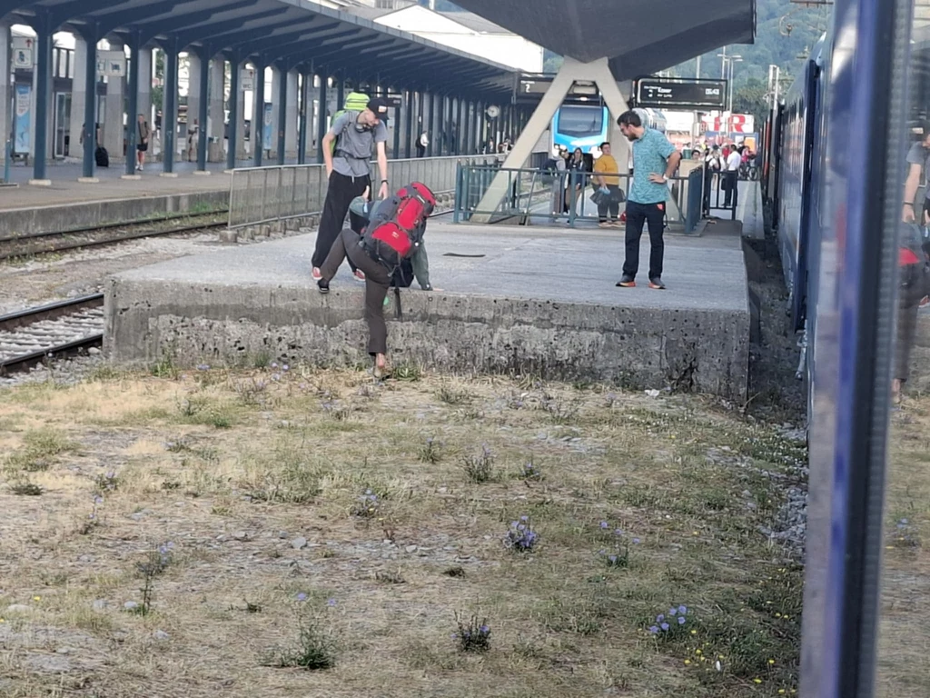Problemy z wysiadaniem w Lublanie Osoba z plecakiem przeskakuje z peronu na ziemię obok torów, w tle widoczne są dwie inne osoby, pociąg oraz zadaszony peron kolejowy.