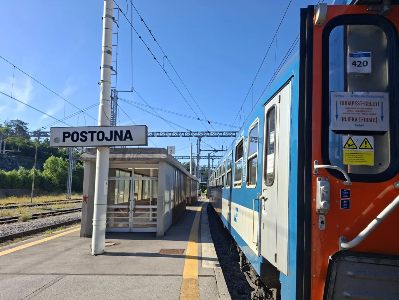 Peron stacji kolejowej Postojna z tablicą z nazwą miasta, na pierwszym planie fragment niebiesko-pomarańczowego wagonu pociągu, po lewej stronie zadaszona wiata oraz słupy trakcyjne i przewody elektryczne.