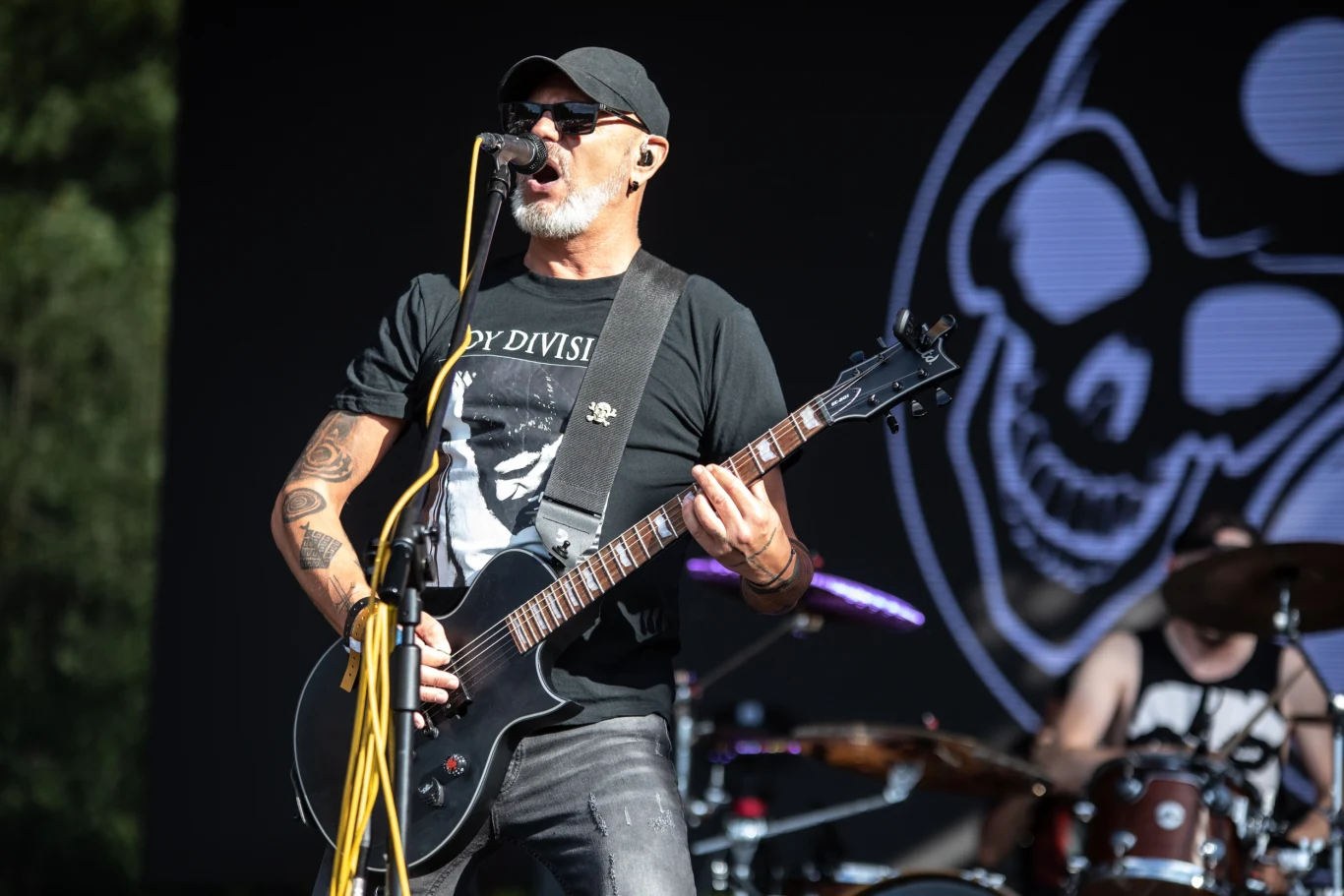 Muzyk grający na gitarze elektrycznej i śpiewający do mikrofonu podczas koncertu na scenie plenerowej, w tle widoczny perkusista oraz fragment dużej grafiki z czaszką.