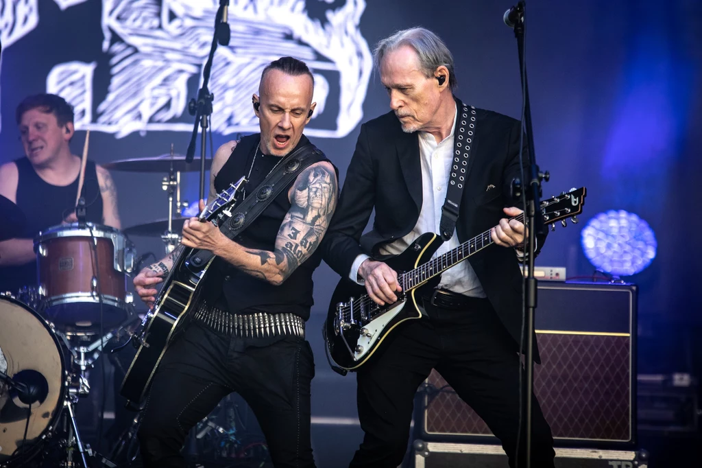 Nergal i John Porter w składzie Me And That Man Dwóch gitarzystów energetycznie gra na gitarach elektrycznych na scenie koncertowej, w tle widoczny perkusista i elementy scenografii oraz sprzęt muzyczny, silne światła reflektorów podkreślają atmosferę występu na żywo.