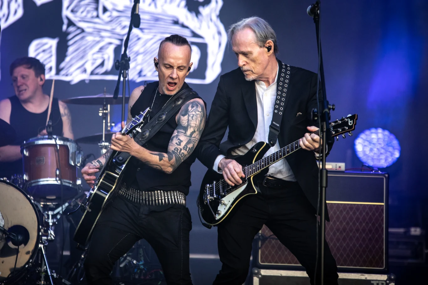 Dwóch gitarzystów energetycznie gra na gitarach elektrycznych na scenie koncertowej, w tle widoczny perkusista i elementy scenografii oraz sprzęt muzyczny, silne światła reflektorów podkreślają atmosferę występu na żywo.