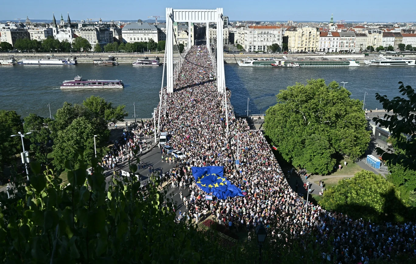 Tłumy na paradzie równości w Budapeszcie. Viktor Orban zabrał głos Tłum ludzi maszerujący przez most nad rzeką, widoczna duża niebieska flaga Unii Europejskiej niesiona przez uczestników, w tle panorama miasta z budynkami oraz łodzie na wodzie.