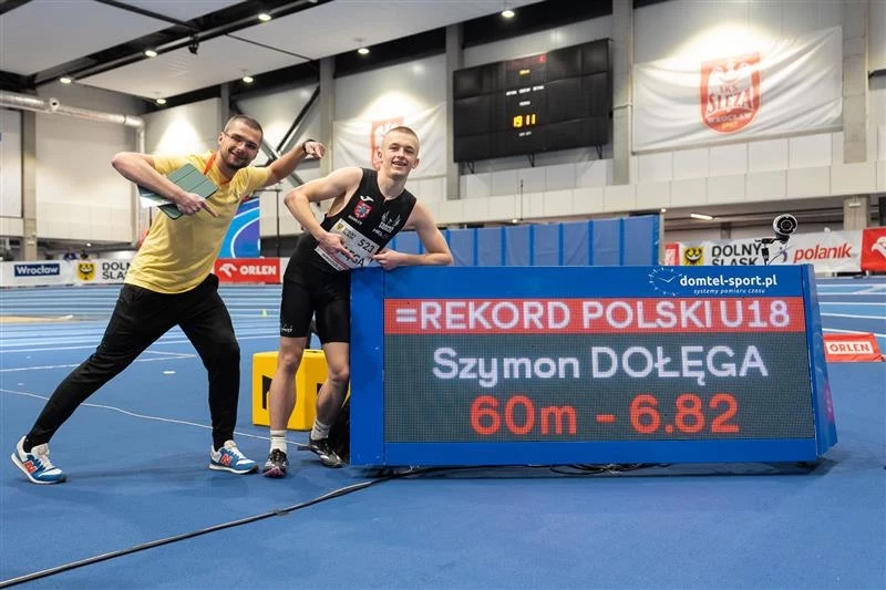 Dwóch młodych mężczyzn pozuje na hali lekkoatletycznej obok elektronicznej tablicy wyświetlającej informację o rekordu Polski U18 na 60 metrów z czasem 6,82 sekundy ustanowionym przez Szymona Dołęgę