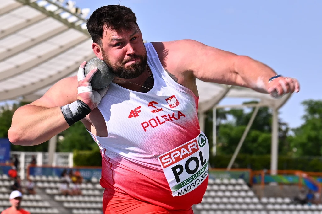 Konrad Bukowiecki Zawodnik w narodowym stroju Polski wykonuje rzut kulą na stadionie lekkoatletycznym, w tle widoczne trybuny i zadaszenie areny sportowej.