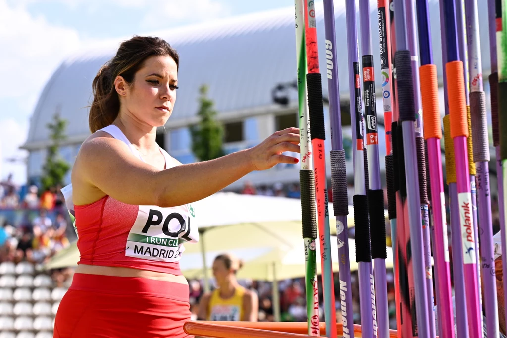 Maria Andrejczyk Zawodniczka w stroju sportowym wybiera tyczkę do skoku o tyczce podczas zawodów lekkoatletycznych na stadionie, w tle widoczna publiczność oraz inne zawodniczki.