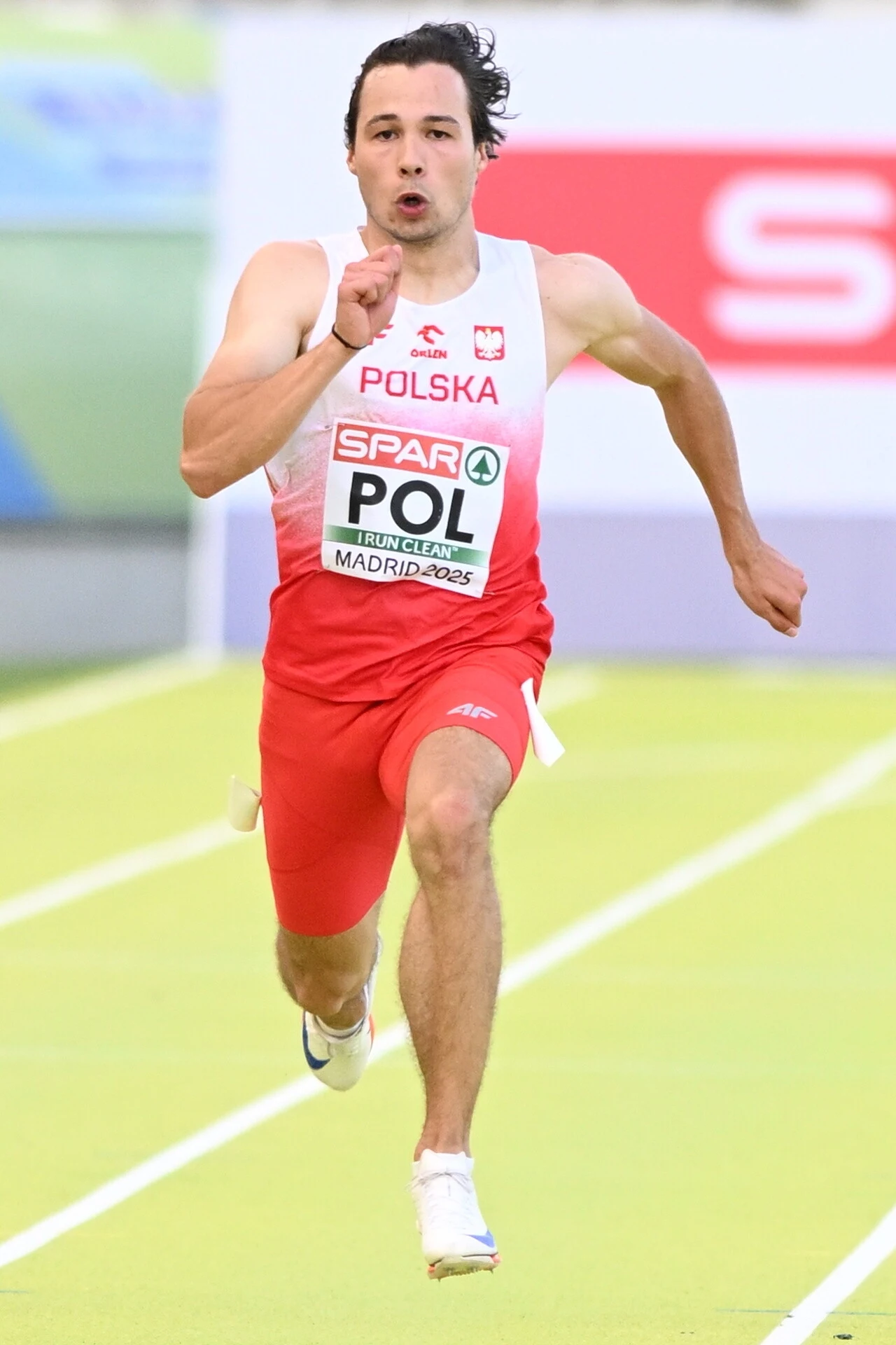 Igor Bogaczyński Lekkoatleta w czerwono-białym stroju reprezentacji Polski biegnie po bieżni podczas zawodów lekkoatletycznych, intensywna ekspresja na twarzy, dynamiczna poza.