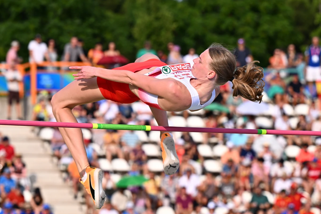 Maria Żodzik Lekkoatletka w biało-czerwonym stroju wykonuje skok wzwyż nad poprzeczką podczas zawodów na stadionie, w tle widoczna publiczność na trybunach.