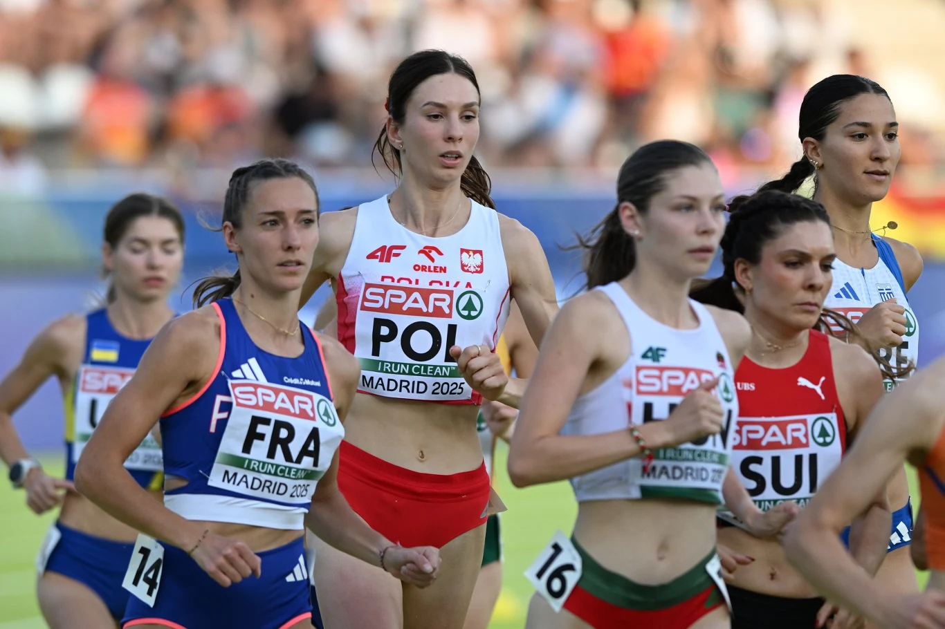 Grupa lekkoatletek biegnących w trakcie zawodów na stadionie, zawodniczki reprezentują różne kraje i mają widoczne numery startowe oraz stroje narodowe.