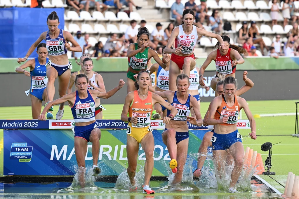 Kinga Krółik nad rowem nad wodą Grupa zawodniczek z różnych krajów pokonuje przeszkodę wodną podczas biegu na stadionie lekkoatletycznym, w tle trybuny z widownią i banery promujące zawody.
