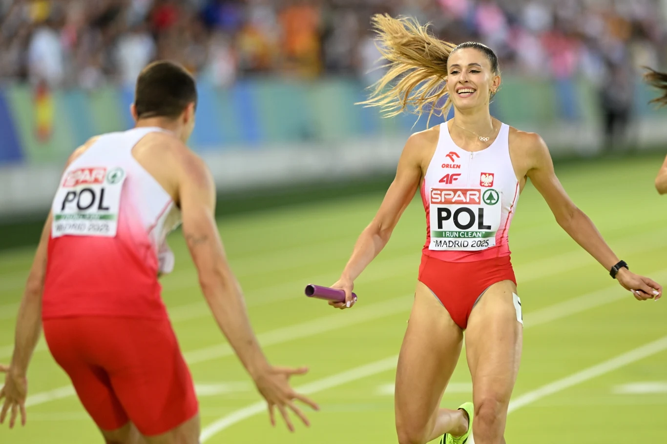 Natalia Bukowiecka na mecie. Tyłem Daniel Sołtysiak Zawodniczka reprezentacji Polski z pałeczką biegnie w kierunku członka swojej drużyny, ubrana w biało-czerwony strój startowy, na stadionie podczas sztafety lekkoatletycznej w otoczeniu publiczności.