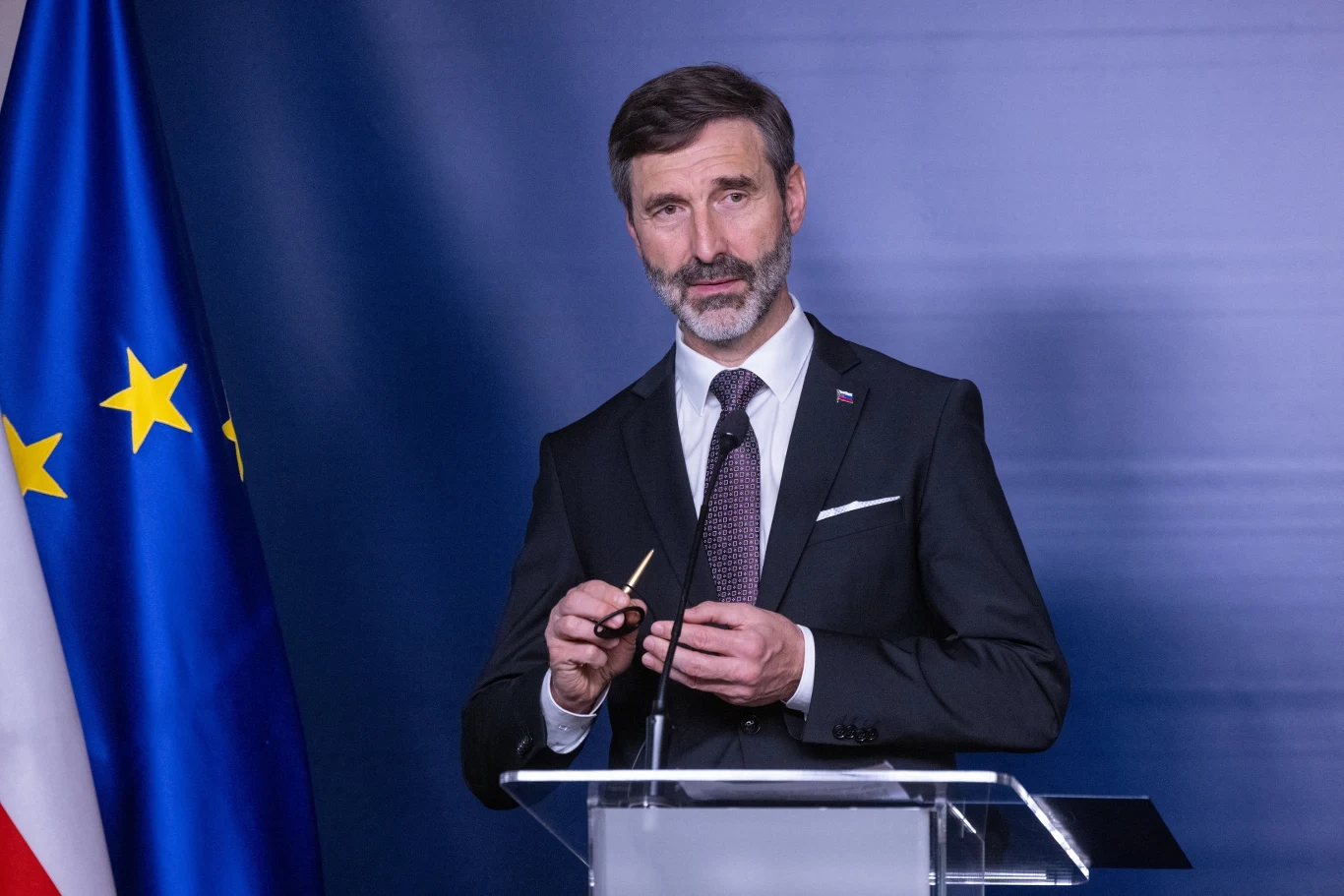 Juraj Blanar sugeruje, że być może należy przebaczyć Rosji Mężczyzna w garniturze przemawiający przy mównicy, trzymający okulary w dłoniach, w tle flaga Unii Europejskiej oraz biało-czerwona flaga narodowa.