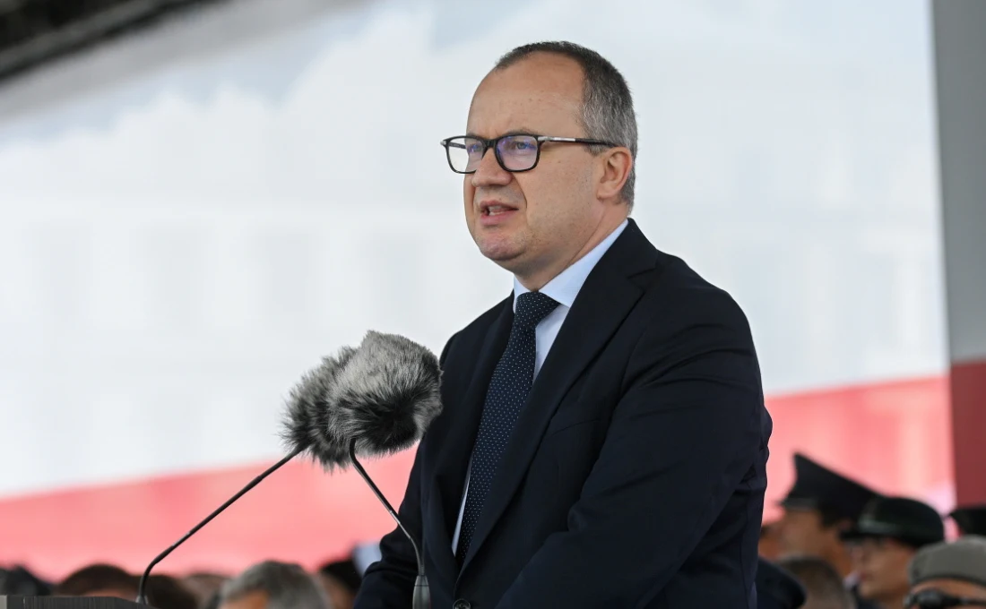 Minister sprawiedliwości i prokurator generalny Adam Bodnar Mężczyzna w garniturze i okularach przemawia do mikrofonu na tle dużej biało-czerwonej flagi, obok widoczni są mundurowi siedzący w cieniu.