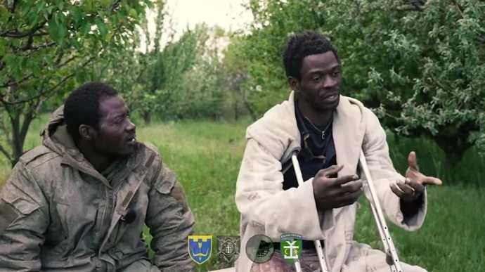 Ukraina. Dwóch obywateli Kamerunu zostało siłą wcielonych do rosyjskiej armii Dwóch mężczyzn siedzących na trawie wśród drzew, jeden z nich ma na sobie jasny płaszcz i opiera się na kulach, obaj wyglądają na zamyślonych i prowadzą rozmowę.