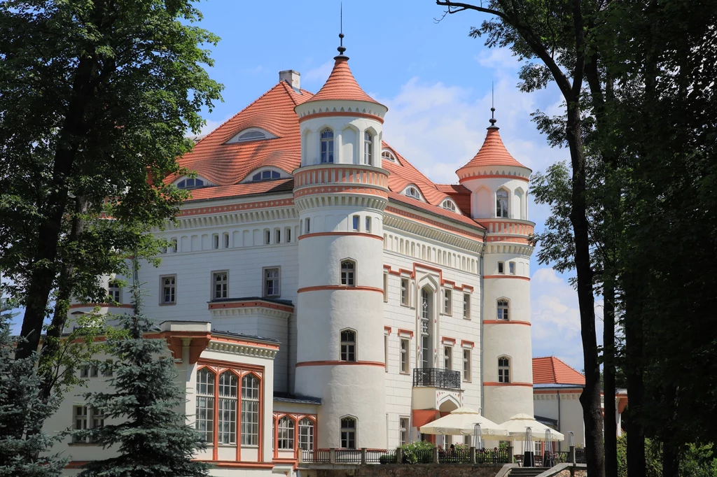 Pałac Wojanów. Elegancki biały pałac z czerwonym dachem i wieżyczkami wyłania się spośród drzew na tle błękitnego nieba, na tarasie widoczne są parasole i stoliki.