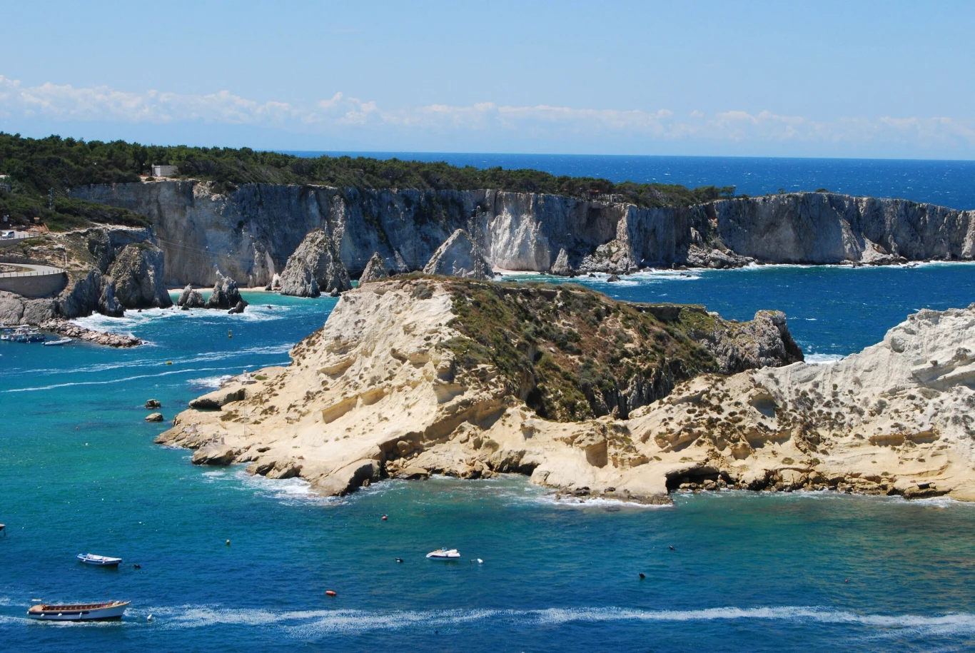 Skały o jasnej barwie otoczone turkusową wodą, na horyzoncie klify porośnięte lasem sosnowym, kilka łodzi unoszących się na spokojnym morzu, bezchmurne niebo.