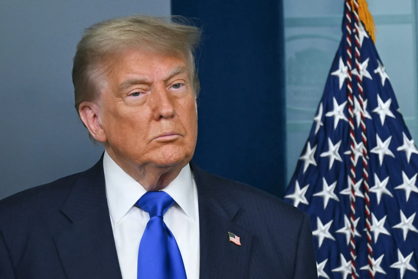 Donald Trump powiedział ile mogą potrwać ataki na Iran Mężczyzna w garniturze z niebieskim krawatem stoi przed flagą Stanów Zjednoczonych, w tle widoczny zarys okna lub plakatu.