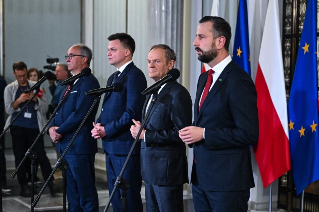 Czterech mężczyzn w garniturach stoi przy mikrofonach podczas konferencji prasowej lub oficjalnego wystąpienia, w tle widoczne flagi Polski i Unii Europejskiej.
