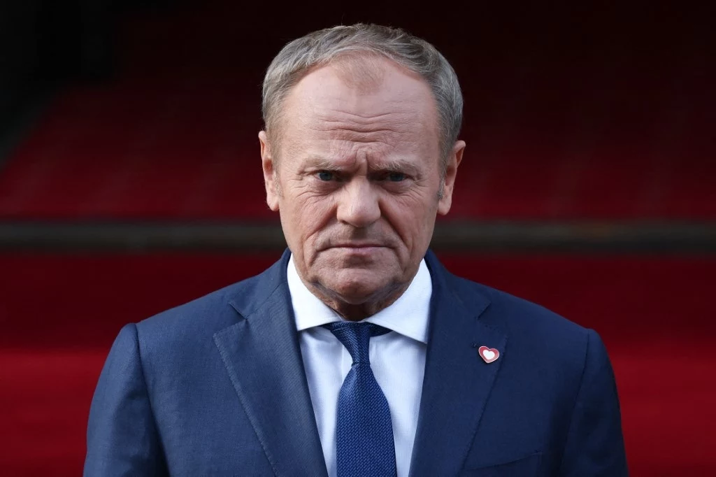 Premier Polski Donald Tusk Mężczyzna w średnim wieku ubrany w granatowy garnitur i niebieski krawat, stoi na tle czerwonych schodów, na klapie marynarki przypięte serduszko WOŚP.