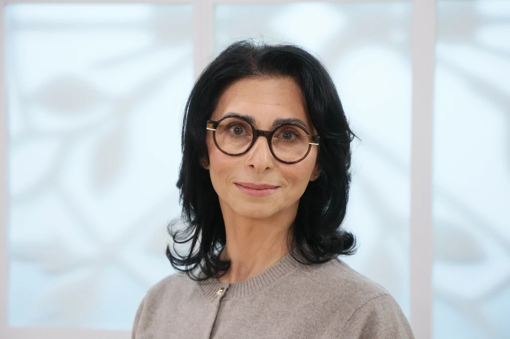 Aida Kosojan-Przybysz Kobieta o ciemnych włosach, nosząca okulary w okrągłych oprawkach oraz beżowy sweter stoi na tle jasnego, rozmytego okna z delikatnym wzorem.