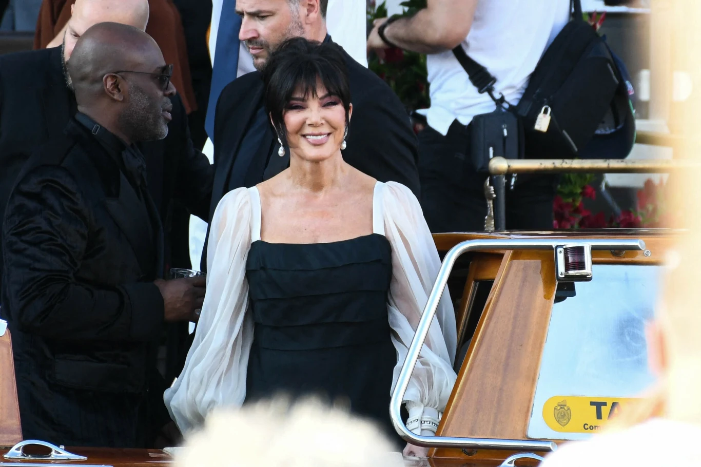 Kris Jenner zachwyciła w Wenecji Elegancko ubrana kobieta z ciemnymi włosami, w czarnej sukni z jasnymi, przezroczystymi rękawami, stoi obok drewnianej łodzi typu taxi wodne, wśród grupy dorosłych osób podczas oficjalnego wydarzenia.