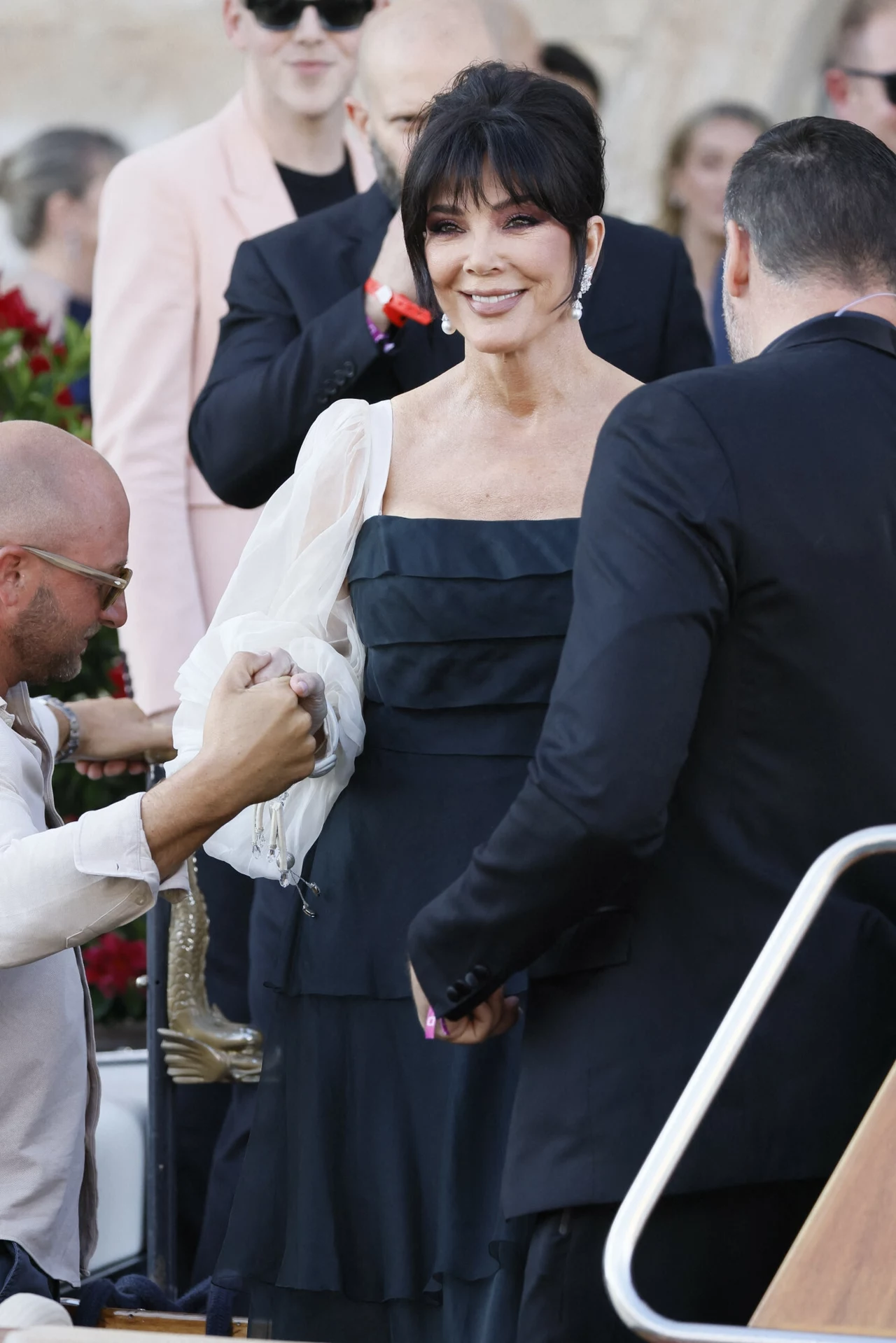Prócz kreacji wzrok przyciągała także fryzura Jenner i jej odświeżona twarz Elegancko ubrana kobieta w czarnej sukience z modnymi bufiastymi rękawami stoi wśród osób ubranych formalnie, jedna z osób podaje jej rękę na powitanie lub w geście pomocy.