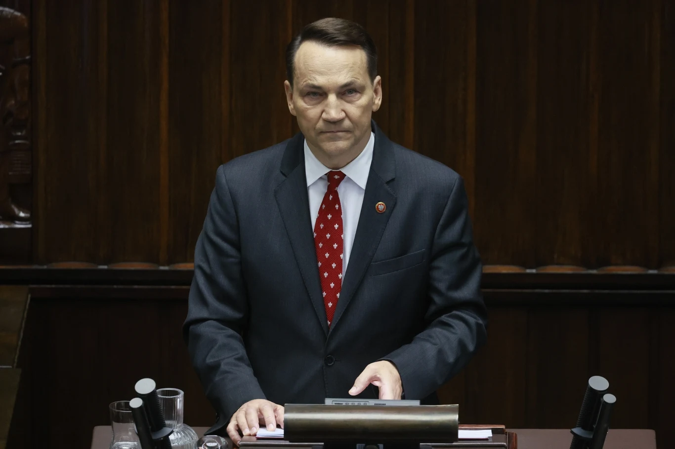 Szef MSZ Radosław Sikorski Mężczyzna w garniturze i czerwonym krawacie przemawia zza mównicy, na której znajdują się dokumenty i mikrofony, w tle widoczne drewniane elementy sali.