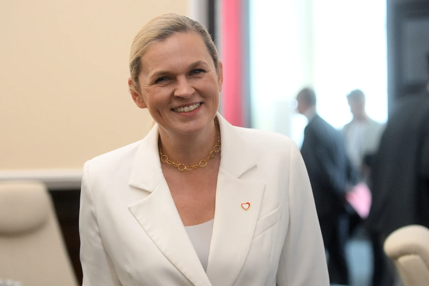 Barbara Nowacka Kobieta w białej marynarce z przypinką w kształcie serca, uśmiechnięta, stoi w pomieszczeniu urzędowym, w tle niewyraźne sylwetki innych osób.