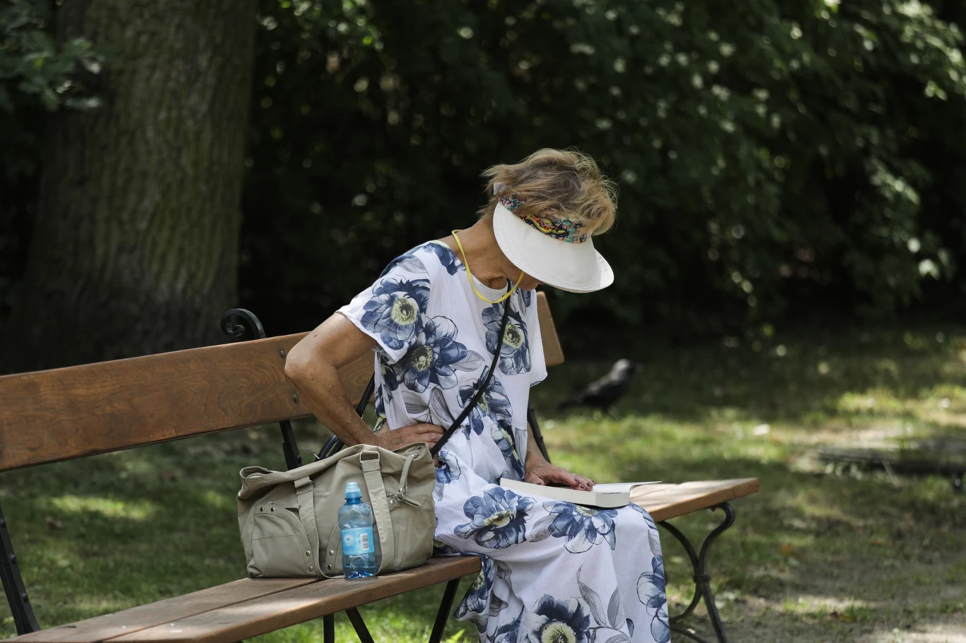 Kto nie otrzyma czternastki? Zdj. ilustracyjne Starsza kobieta w letniej sukience i kapeluszu z szerokim rondem siedzi na ławce w parku i czyta książkę, obok niej jasna torebka oraz butelka wody mineralnej, w tle drzewa i zieleń.
