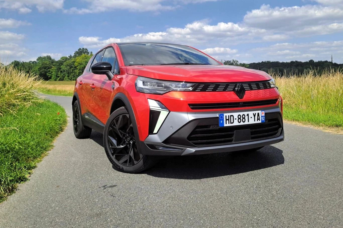 Czerwony samochód osobowy typu SUV marki Opel stoi zaparkowany na wiejskiej, asfaltowej drodze otoczonej wysoką trawą i zielenią, pod jasnym niebem z pojedynczymi chmurami.