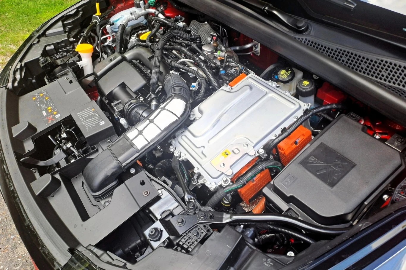 Komora silnika samochodu hybrydowego z wyraźnie widocznymi elementami układu napędowego, przewodami, pokrywą silnika oraz osprzętem technicznym, w tym złącza oznaczone charakterystycznymi pomarańczowymi osłonami dla wysokiego napięcia.