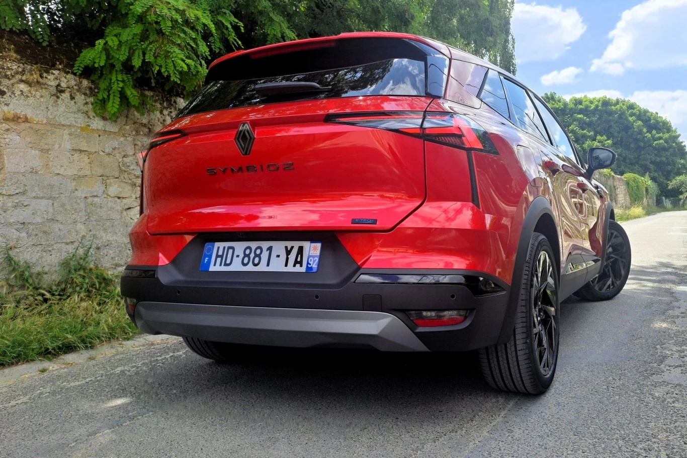 Czerwony samochód marki Renault Symbioz zaparkowany na poboczu asfaltowej drogi, otoczony zielenią i kamiennym murem, widok od tyłu ukazujący tablicę rejestracyjną i nowoczesny design auta.