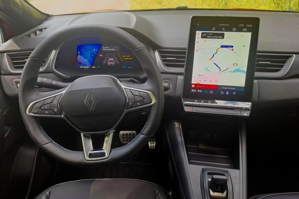 Wewnątrz Renault Symbioz w większości nie uległo zmianom. Od razu jednak zauważymy nową dźwignię zmiany biegów, znaną choćby z Dacii Spring. Kokpit nowoczesnego samochodu z cyfrowym ekranem między zegarami oraz dużym, dotykowym panelem centralnym wyświetlającym mapę nawigacji. Widoczna kierownica z logo marki oraz minimalistyczny design wnętrza.