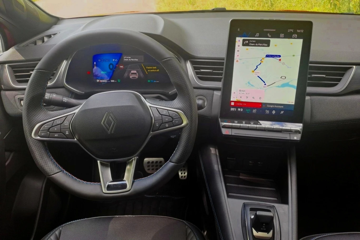 Kokpit nowoczesnego samochodu z cyfrowym ekranem między zegarami oraz dużym, dotykowym panelem centralnym wyświetlającym mapę nawigacji. Widoczna kierownica z logo marki oraz minimalistyczny design wnętrza.