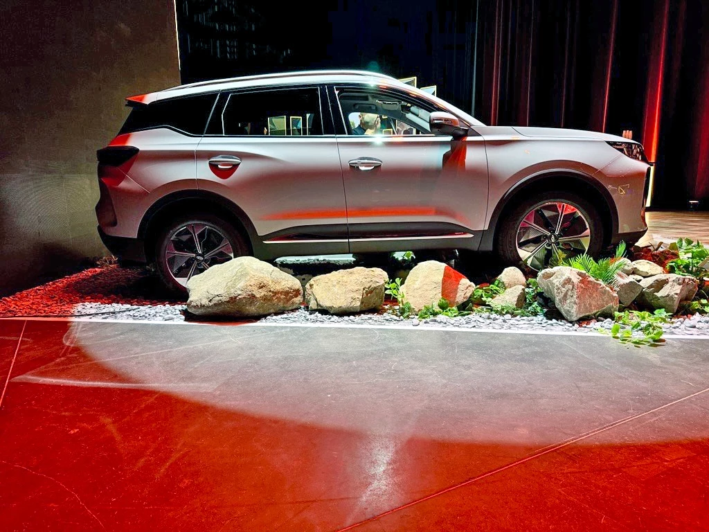 Chery Tiggo 7 Nowoczesny SUV w kolorze srebrnym zaparkowany na ekspozycji, wokół ustawiono ozdobne kamienie i roślinność, auto o dynamicznych liniach i dużych felgach.
