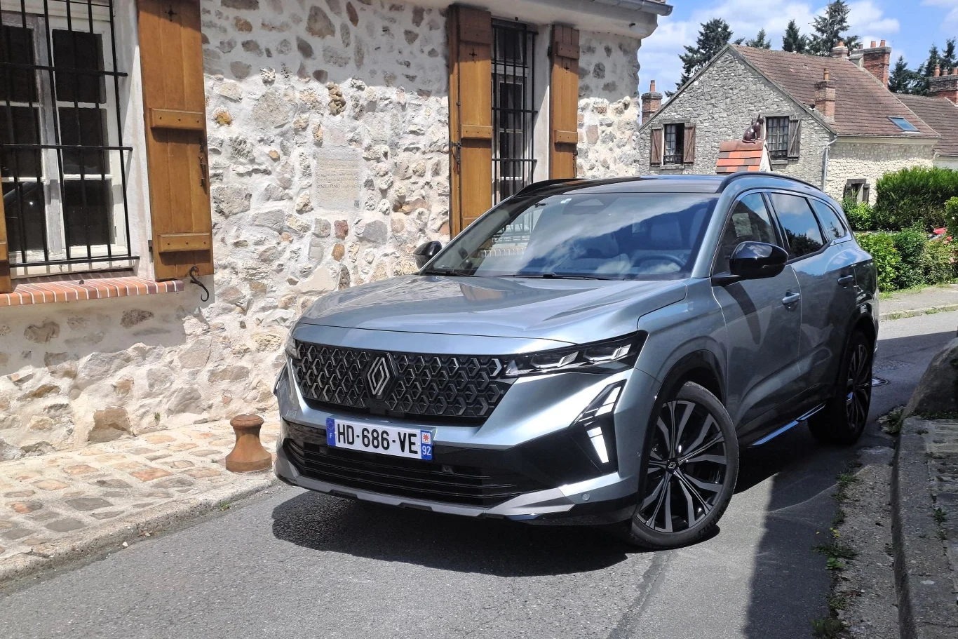 Srebrny nowoczesny SUV zaparkowany przy zabytkowym budynku z kamienia, z zamkniętymi drewnianymi okiennicami, na wąskiej uliczce w spokojnej, wiejskiej okolicy.