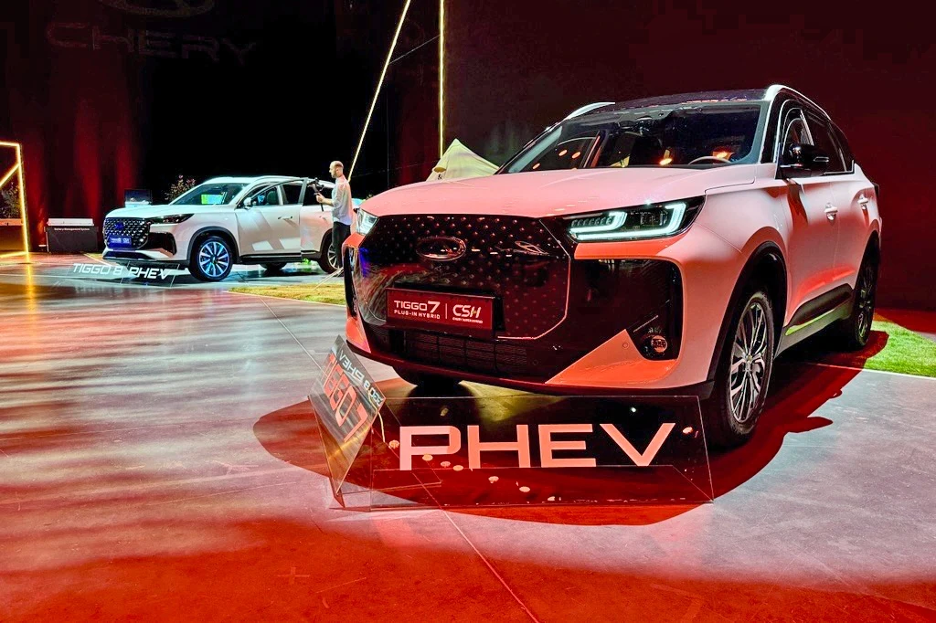 Dwa nowoczesne SUV-y marki Chery, modele Tiggo 7 i CS1, eksponowane w jasnym oświetleniu na tle ciemnej hali wystawowej, podkreślone napisem PHEV oraz informacjami o wersji hybrydowej plug-in