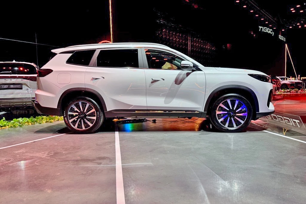 Chery Tiggo 8 Biały SUV klasy średniej ustawiony bokiem na ekspozycji, wyeksponowane nowoczesne linie nadwozia, duże aluminiowe felgi oraz przyciemniane szyby, pojazd prezentowany w oświetlonym wnętrzu hali targowej.