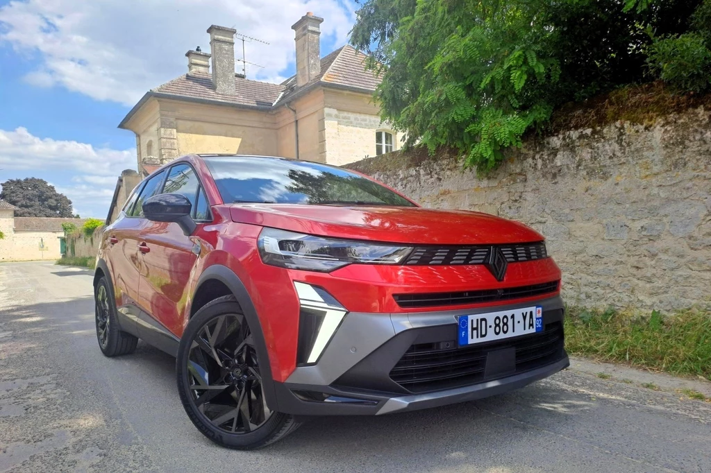 Renault deklaruje, że Symbioz w tej wersji napędowej oferuje zasięg do 1 tys. km. Czerwony samochód osobowy zaparkowany na wąskiej, miejskiej ulicy, w tle kamienny mur oraz zabytkowy budynek z jasnego kamienia, po lewej stronie widoczna roślinność.