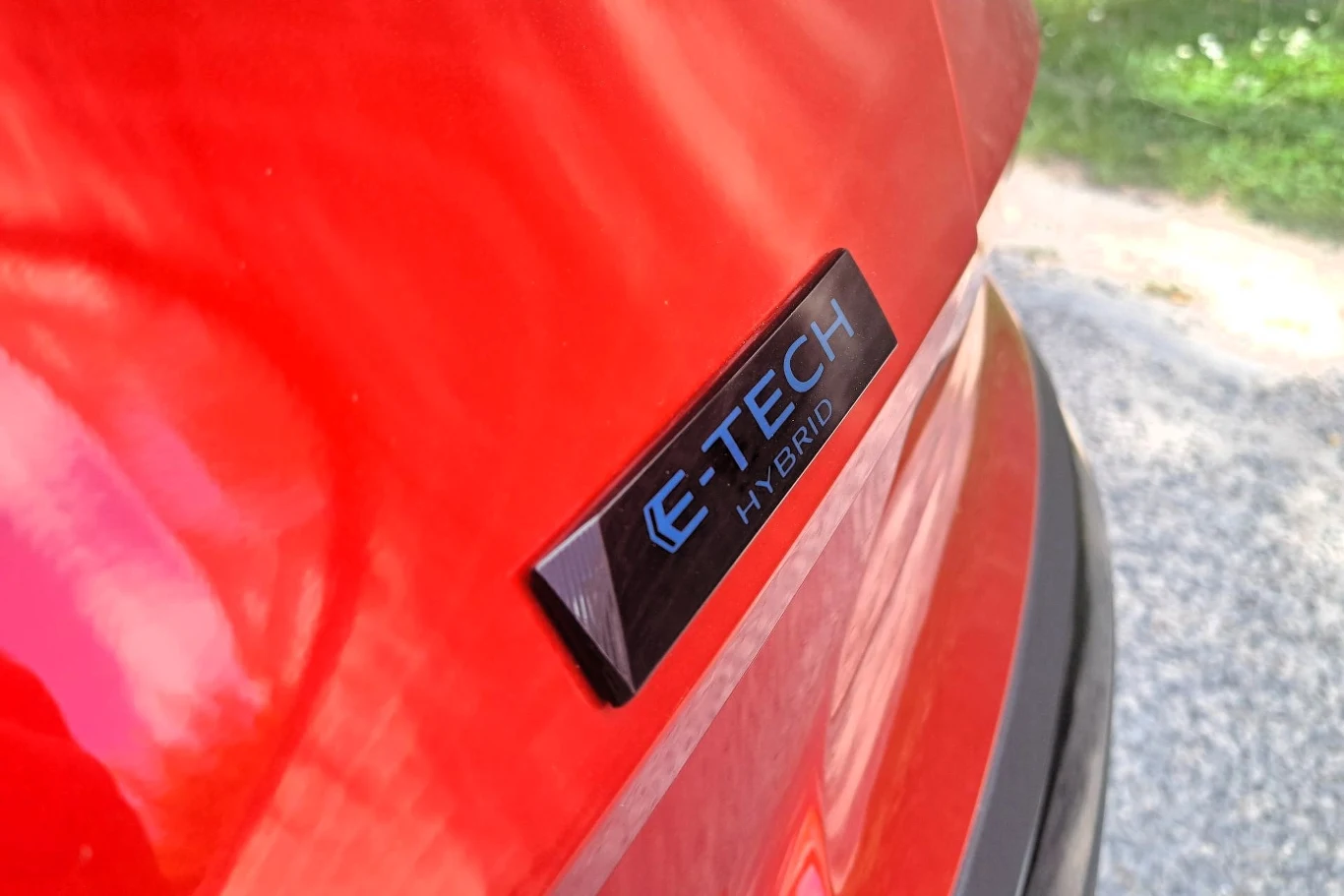 Czerwony błotnik samochodu z wyraźnym znakiem E-TECH HYBRID na tle żwirowej nawierzchni i fragmentem zieleni.