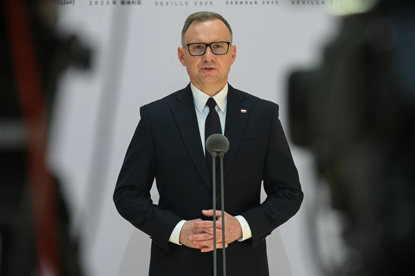 Prezydent Andrzej Duda ocenił ruch Adama Bodnara Mężczyzna w garniturze i okularach stoi za mównicą, trzymając ręce złożone, patrzy prosto w obiektyw, w tle niewyraźne sylwetki kamer.