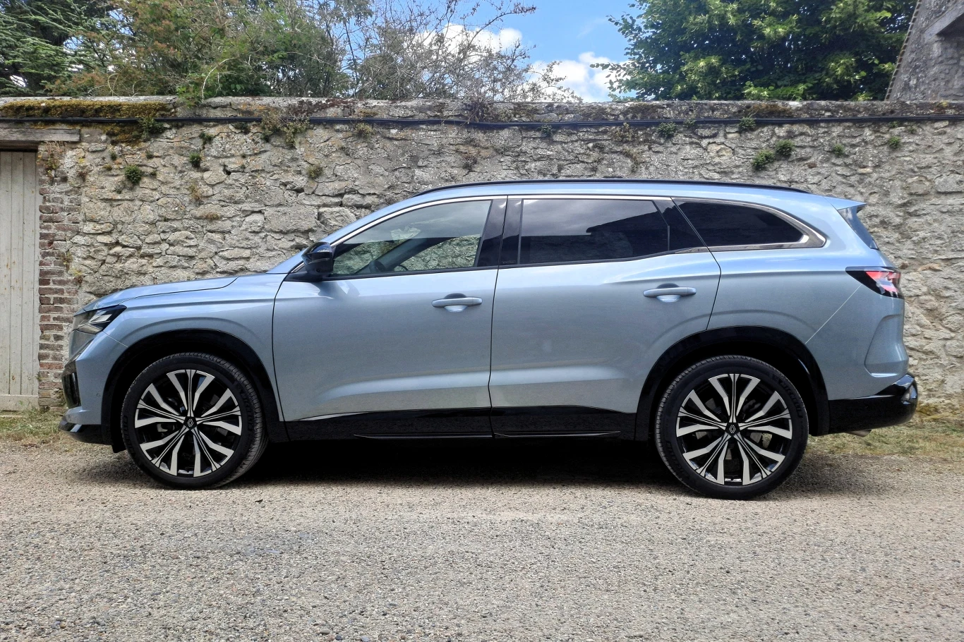 Srebrny SUV nowoczesnej konstrukcji zaparkowany na szutrowej drodze, w tle widoczny stary kamienny mur oraz fragmenty zieleni i nieba.