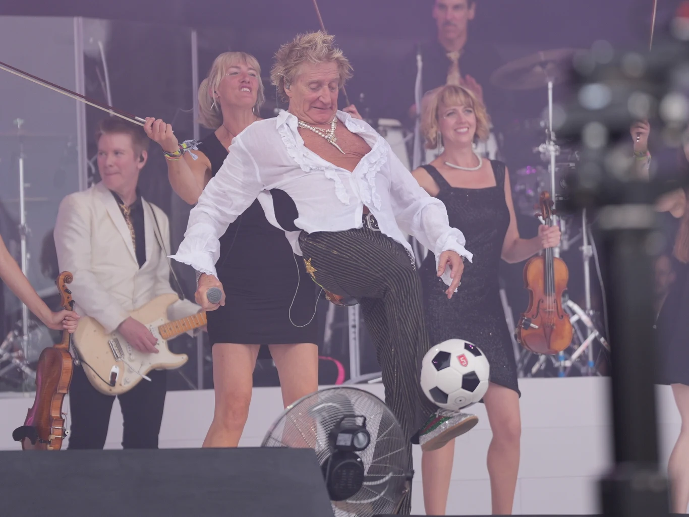 Rod Stewart podczas Glastonbury Festival 2025 Mężczyzna w rozpiętej koszuli kopiący piłkę nożną na scenie, otoczony przez uśmiechniętych muzyków grających na różnych instrumentach, w tym gitarze i skrzypcach, w tle sceniczny sprzęt muzyczny.