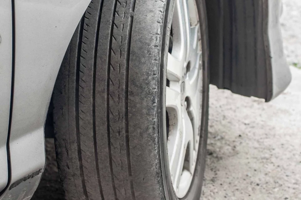 Zużyta opona samochodowa zamontowana na feldze, z wyraźnymi śladami starcia bieżnika, pokazana z bliska na tle asfaltu.