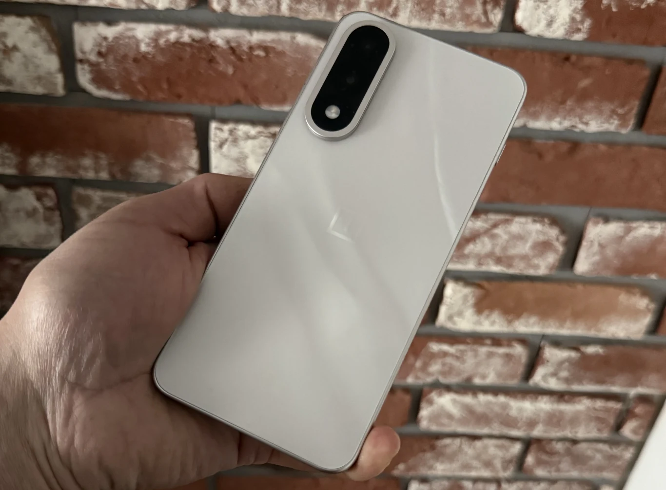 Test OnePlus Nord 5. Niedrogi, a mocy mu nie brakuje/ Biała obudowa smartfona trzymanego w lewej dłoni, na tle czerwonej ceglanej ściany, widoczny podłużny czarny moduł aparatu z dwoma obiektywami i lampą błyskową.