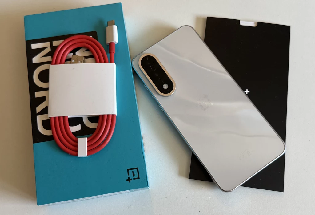 Zawartość opakowania smartfona OnePlus Nord 5. Zestaw jest podstawowy. Nowoczesny smartfon z białą obudową, czerwony kabel USB-C, niebieskie pudełko z logo OnePlus Nord oraz czarny element opakowania, wszystko ułożone na białym tle.