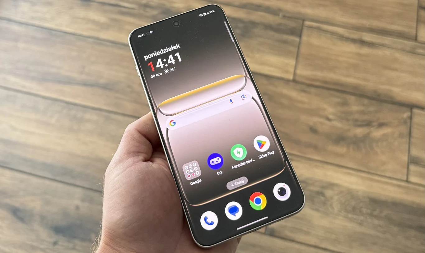 OnePlus Nord 5 ma jasny i czytelny wyświetlacz. Osoba trzyma w dłoni nowoczesny smartfon z wyświetlaczem ukazującym pulpit, na którym widoczne są ikony najpopularniejszych aplikacji oraz panel powiadomień, na tle drewnianej podłogi.