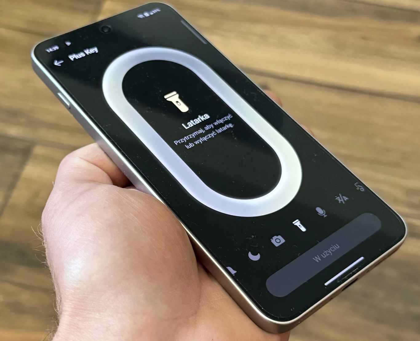 OnePlus Nord 5 wprowadza przycisk Plus Key znany z ostatnich flagowców marki. Smartfon trzymany w dłoni z wyświetlonym ekranem konfiguracji przycisku funkcyjnego, pokazującym możliwość ustawienia latarki na ten przycisk, na tle drewnianej podłogi.